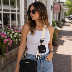 A woman wearing a white tank top and light blue jeans models a black crossbody bag with a cream strap. The bag features a small black pouch at the center of the strap with the brand name 'G128' printed in white.