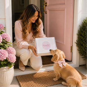 A woman kneels at a pink front door holding a white box labeled 'Dining Gear' while a golden retriever dog wearing a pink bow looks on. Pink flowers and a brown doormat are visible in the scene.