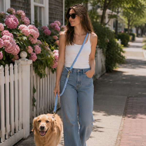 A woman walking a golden retriever on a sidewalk in front of a house with pink hydrangeas. She is wearing a white tank top, light blue wide-leg jeans, white sneakers, and a light blue crossbody bag.
