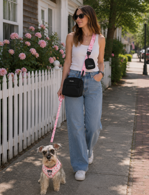 A woman walking a small dog on a pink and white polka dot leash and matching harness on a sunny residential sidewalk. She is wearing a white tank top, light blue jeans, and black crossbody bags.