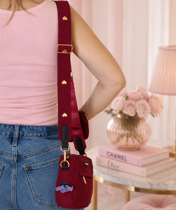A burgundy fabric crossbody bag with a wide matching shoulder strap, gold-tone hardware, and a small purple plush accent, shown worn by a person in a pink ribbed tank top and blue jeans in a softly styled room.