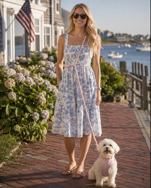 A woman wearing a blue and white toile-patterned midi dress stands on a brick waterfront walkway, holding a leash with a small white dog. Houses, hydrangeas, and water are visible in the background.
