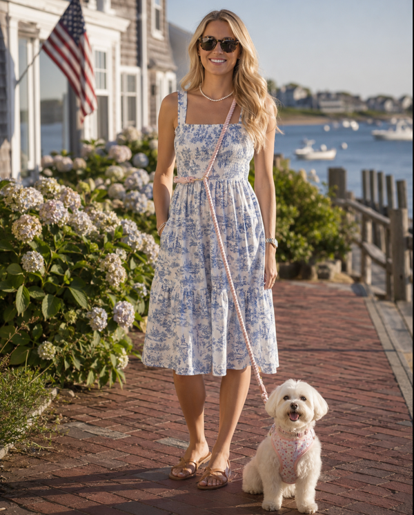 A woman wearing a blue and white toile-patterned midi dress stands on a brick waterfront walkway, holding a leash with a small white dog. Houses, hydrangeas, and water are visible in the background.
