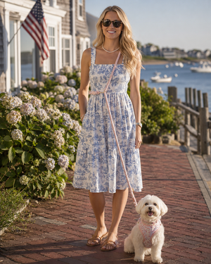 Woman in a blue floral dress walking a white dog on a brick path with a scenic background.