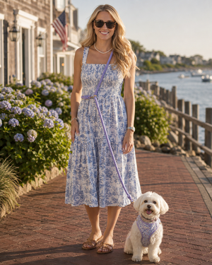Woman in a blue floral dress standing with a white dog on a leash by a waterfront.